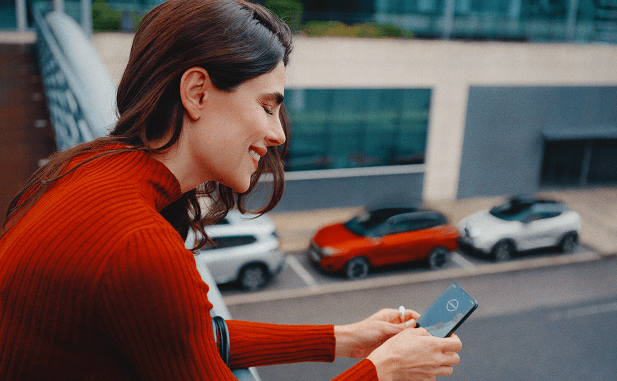 Frau mit rotem Pullover lächelt und schaut auf ihr Smartphone auf einem Balkon mit Blick auf geparkte Nissan Autos.